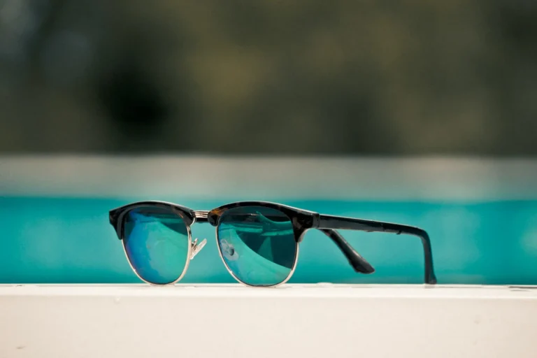 Sunglasses with black frames and reflective blue polarized lenses sit on a white surface next to a blurred, calm turquoise background pool.