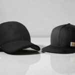 Two black caps on a reflective surface; the left is a curved-brim, and the right is a flat-brim with a logo.