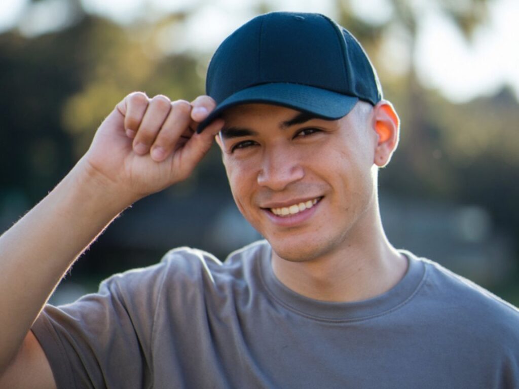 A person smiles warmly while holding the brim of a blue cap outdoors.