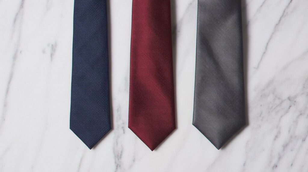 Three neckties of varying widths and lengths in navy blue, burgundy, and gray are neatly arranged on a white marble surface.