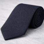 A neatly rolled dark navy Grenadine tie with a subtle textured pattern, resting on a light gray marble surface.