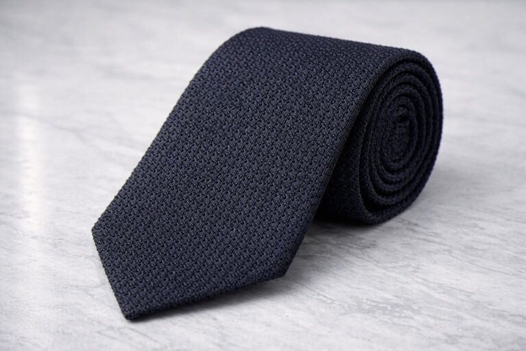 A neatly rolled dark navy Grenadine tie with a subtle textured pattern, resting on a light gray marble surface.