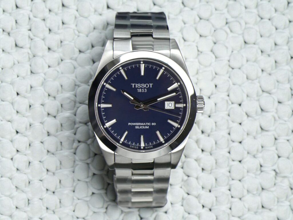 Tissot Gentleman Powermatic 80 Silicium stainless steel watch with a dark blue face, silver hour markers, and a date display, resting on a textured white surface. 