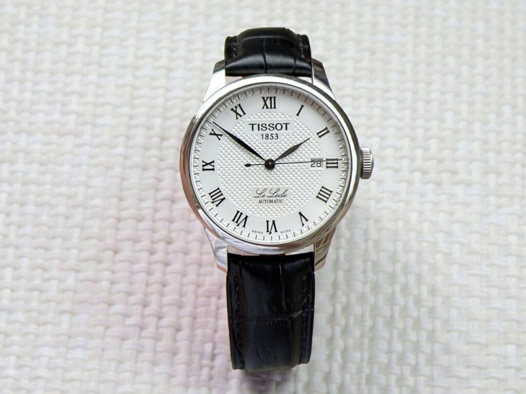 Tissot Le Locle Powermatic 80 watch with a white textured dial, Roman numerals, and a black leather strap, set against a woven, off-white background.