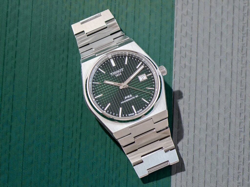 Tissot PRX Powermatic 80 watch with a dark textured dial and date window, resting on a green and gray textured background.