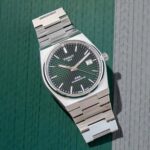 Tissot PRX Powermatic 80 watch with a dark textured dial and date window, resting on a green and gray textured background.