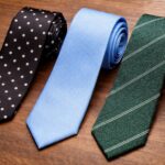 Three budget neckties on a wooden surface: a black tie with white polka dots, a solid sky blue tie, and a green tie with diagonal stripes.