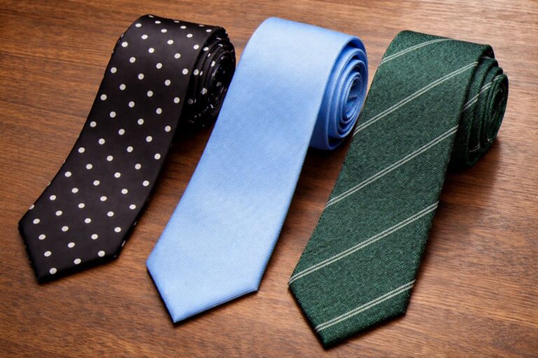 Three budget neckties on a wooden surface: a black tie with white polka dots, a solid sky blue tie, and a green tie with diagonal stripes.