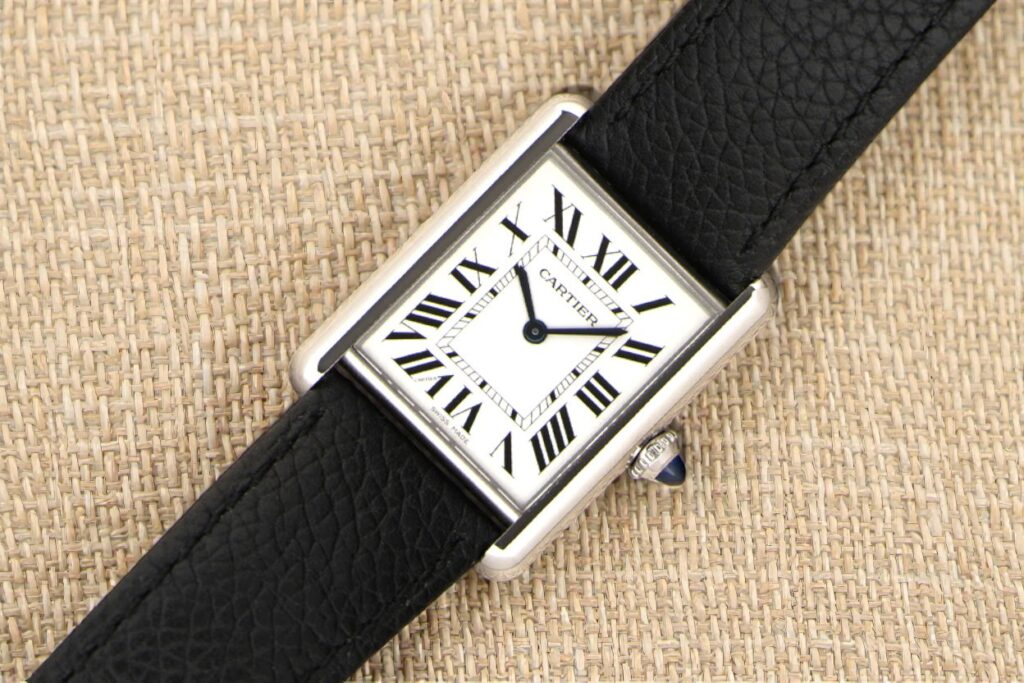 Cartier Tank Must rectangular watch on a textured beige background. Features a black leather strap, a white face with black Roman numerals, and a cabochon crown.