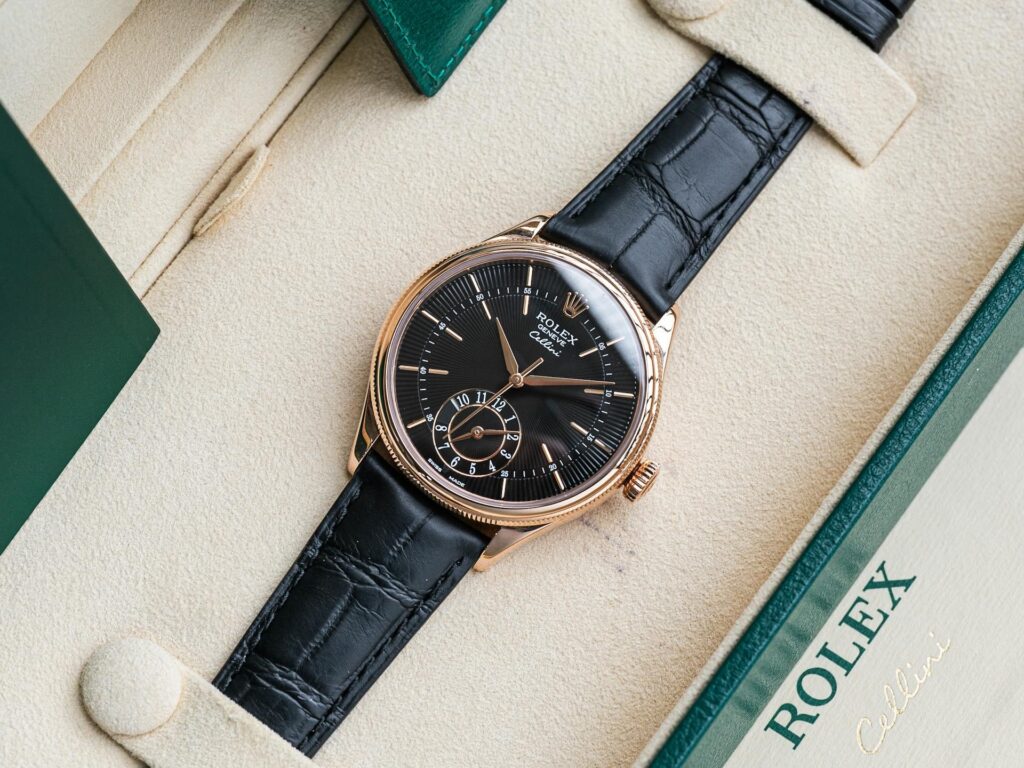 An elegant Rolex Cellini watch with a black leather strap and gold casing rests in a cream box. The dial is black with gold accents and a small seconds subdial.