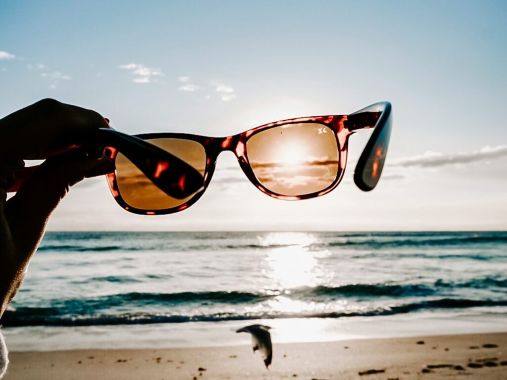 Close-up of a hand holding sunglasses, framing an ocean sunset. Sunlight reflects on the water, creating a warm, serene atmosphere.