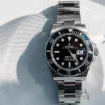 A stainless steel Rolex Submariner Date automatic watch with a black dial and bezel is shown lying on a white surface in natural light.