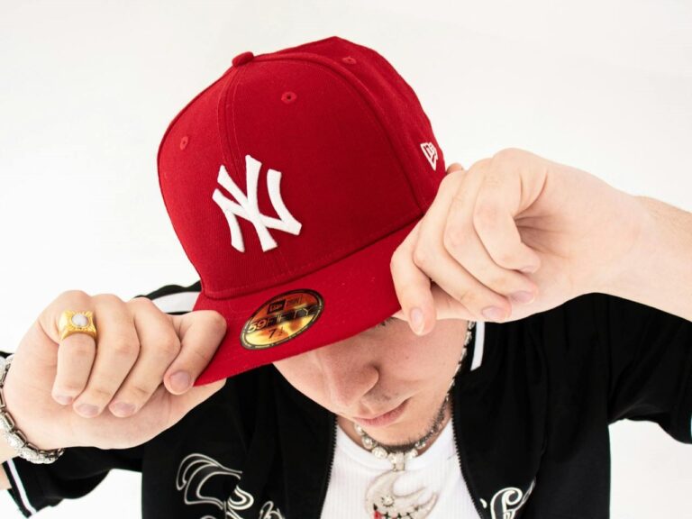 A person tilts a 59Fifty red cap with a white logo, partially covering their face. They wear a black jacket and jewelry, conveying a casual, stylish vibe.