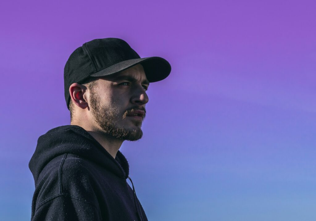A man in a black ball cap and hoodie looks thoughtfully into the distance against a vibrant gradient sky transitioning from purple to blue.