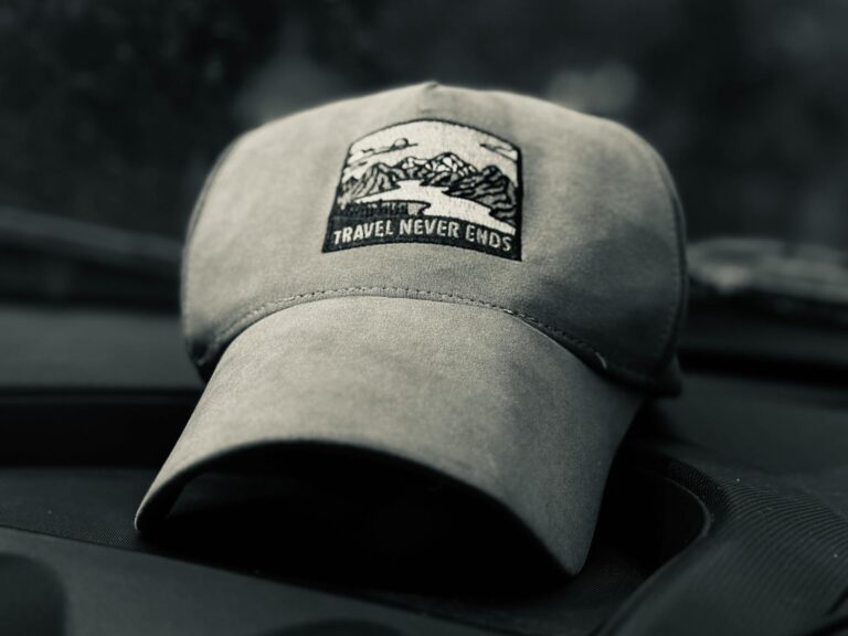Gray baseball cap with a patch featuring mountains and the phrase "Travel Never Ends," resting on a car dashboard.
