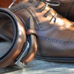 Brown leather dress shoes with detailed stitching are paired with a matching leather belt, elegantly coiled, on a wooden surface.