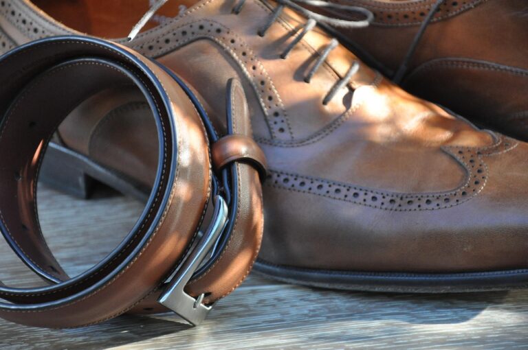 Brown leather dress shoes with detailed stitching are paired with a matching leather belt, elegantly coiled, on a wooden surface.