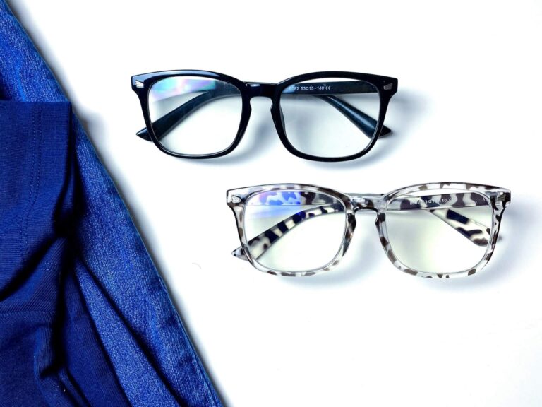 Two pairs of blue light blocking sunglasses on a white surface. One pair is black, the other has a leopard print design. A blue fabric is draped beside them.