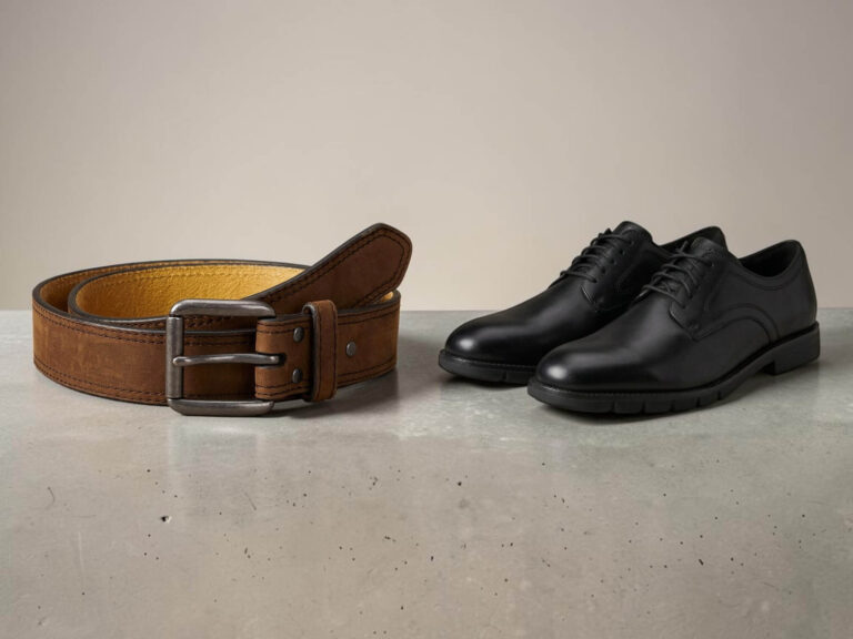 A brown leather belt with a silver buckle is coiled next to a pair of black leather dress shoes on a smooth, gray surface.