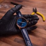 A gloved hand holds a blue-faced wristwatch with a blue leather strap on a wooden table. Nearby are yellow pliers and a watch repair tool.