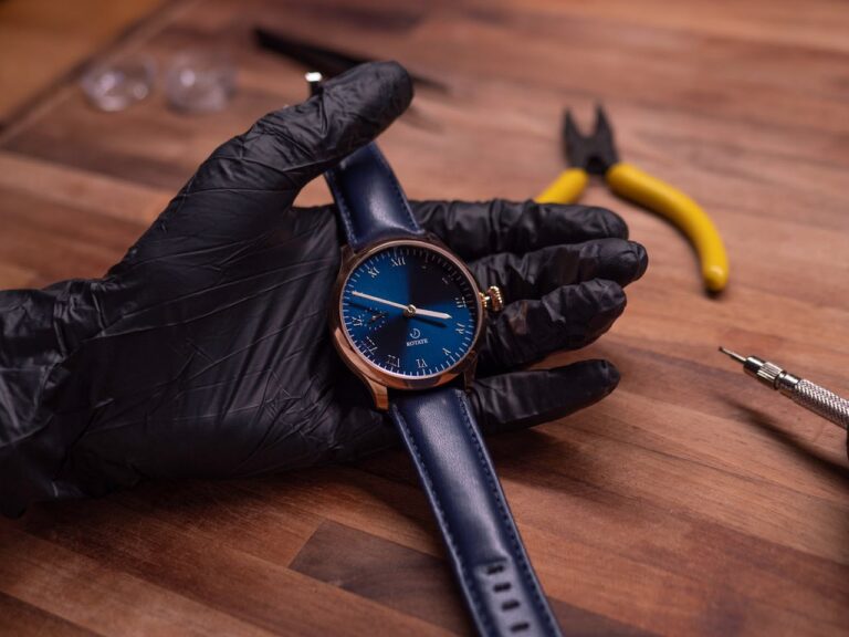 A gloved hand holds a blue-faced wristwatch with a blue leather strap on a wooden table. Nearby are yellow pliers and a watch repair tool.