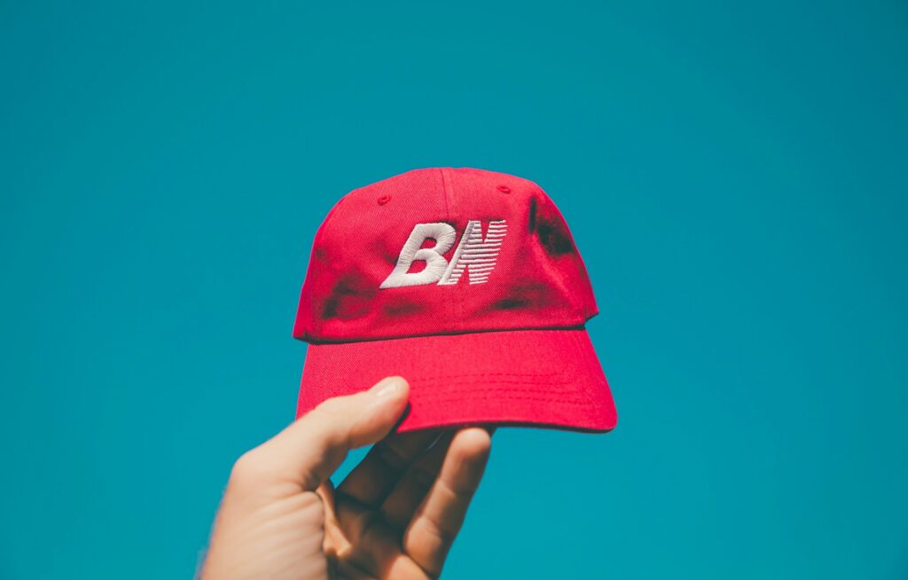 A person holds a red dad cap with 'BN' embroidered in white against a bright blue sky background.