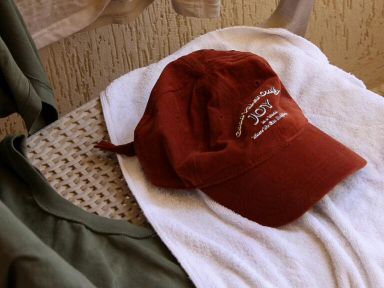 A red baseball cap, freshly washed, with text, lies on a white towel draped over a wicker chair. An olive green shirt is placed nearby.