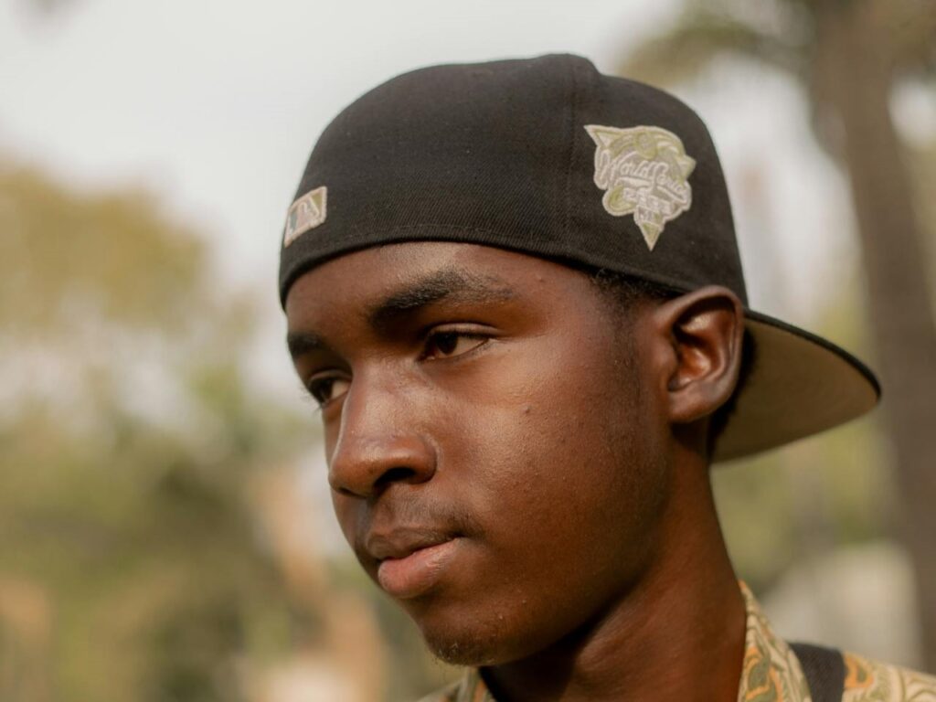 Young man in a sideways black fitted cap looks thoughtful outdoors. The soft-focus background includes trees and greenery.