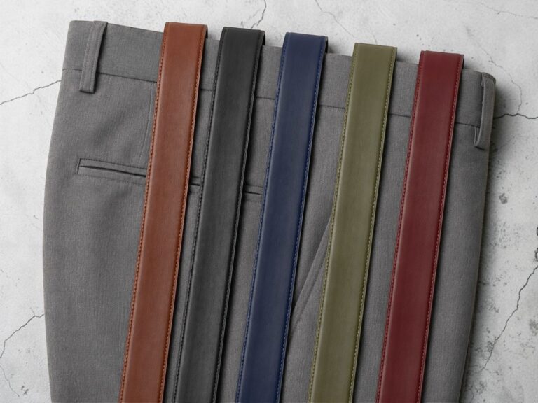 A pair of gray trousers is laid out with five leather belts of different colors: brown, black, blue, olive, and burgundy, arranged neatly across them.