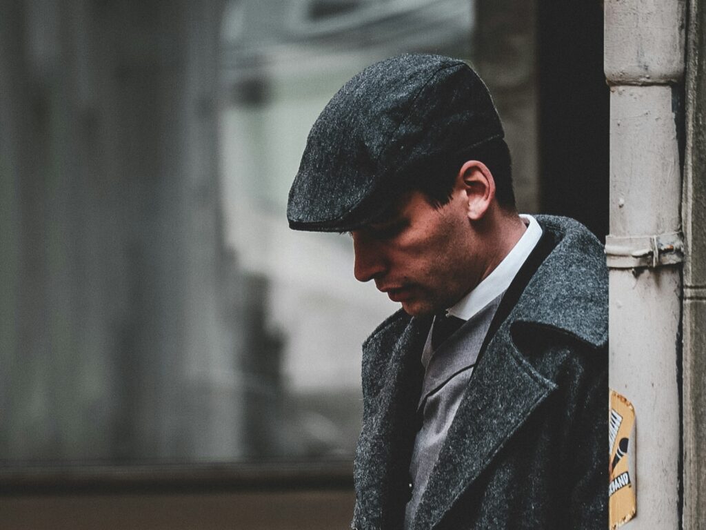 Man in a gray flat cap and coat stands pensively against a wall. The muted tones and soft lighting create a nostalgic, vintage atmosphere.