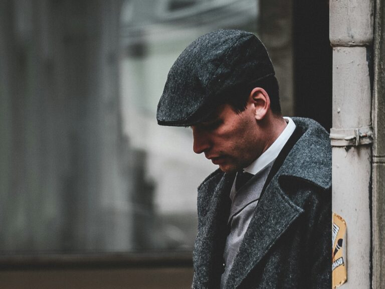Man in a gray flat cap and coat stands pensively against a wall. The muted tones and soft lighting create a nostalgic, vintage atmosphere.