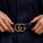 Hands adjusting a black leather designer belt with a gold GG logo buckle, worn over a dark outfit.