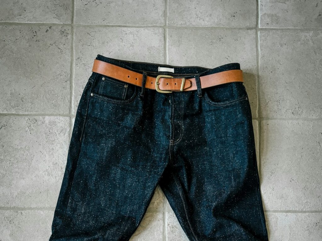 A pair of dark denim jeans with a tan leather belt laid on a light gray tiled floor. 