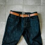 A pair of dark denim jeans with a tan leather belt laid on a light gray tiled floor.