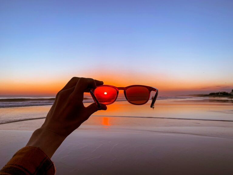 A hand holds polarized sunglasses at a beach sunset, with the sun visible through the lens, casting an orange glow.