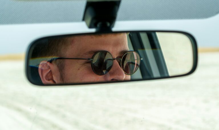 Reflection in a car's rearview mirror showing a man's face wearing round sunglasses, with a calm desert landscape in the background.
