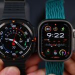 Two smartwatches are held side by side. The left watch has a black strap and circular face displaying analog time. The right has a blue strap and digital interface.