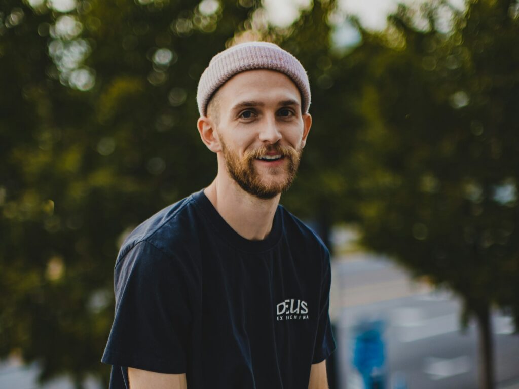 A smiling man with a beard wears a pink knit skull cap and a dark T-shirt outdoors. 