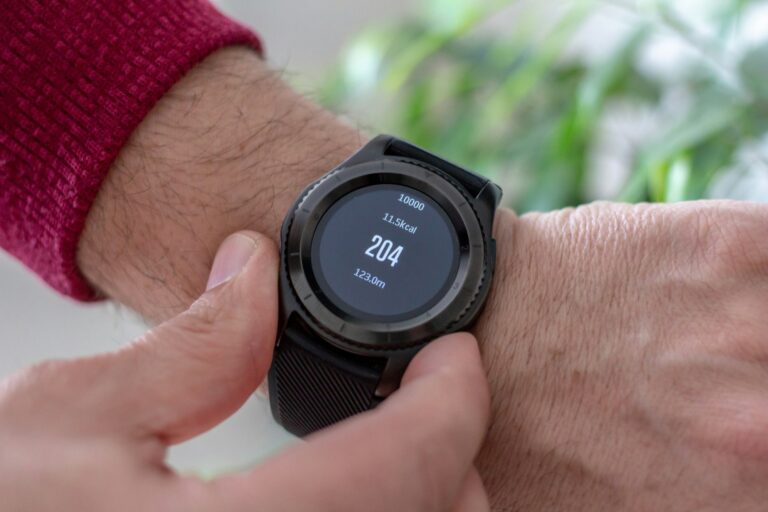 A person wearing a black smartwatch displays fitness stats: 204 steps, 11.5 kcal burned. Finger taps screen. Background has blurred green plants.