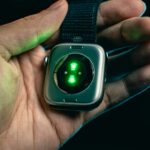 Close-up of a hand holding a smartwatch with a glowing green sensor on the back, set against a dark, blurred background.