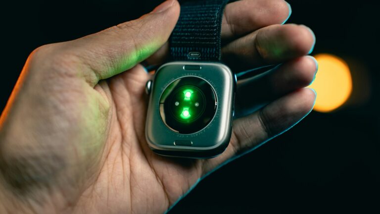 Close-up of a hand holding a smartwatch with a glowing green sensor on the back, set against a dark, blurred background.