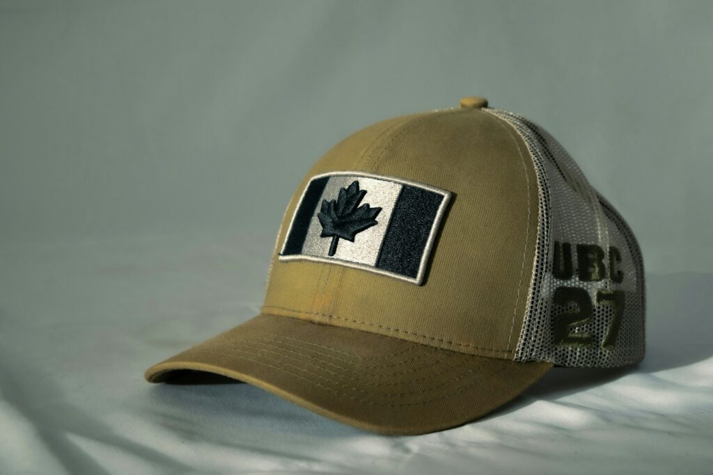 Beige trucker cap with embroidered patch of a black maple leaf on a striped background. "UBC 27" is printed on the mesh panel.