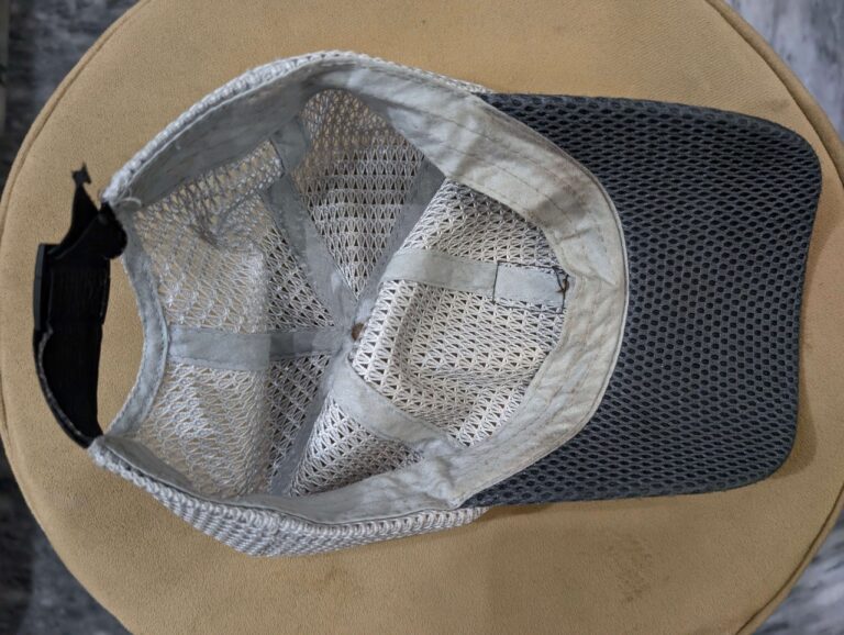 Top view of a beige mesh cap with a black brim on a tan surface. The cap's inside shows ventilation and worn fabric, marked by sweat strain, suggesting frequent use.