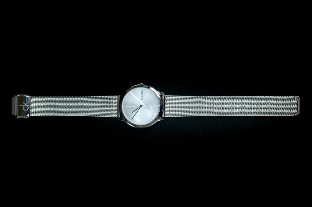 A silver mesh band wristwatch with a minimalist white face and thin black hands is displayed horizontally against a black background.