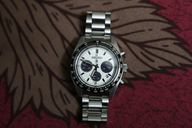 A silver Seiko chronograph watch with a Tachymeter bezel, white dial and black subdials is displayed on a textured red and brown leafy-patterned background.