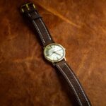 A vintage wristwatch with a gold case and white dial, featuring a brown leather strap with white stitching, lies on a textured brown leather surface.