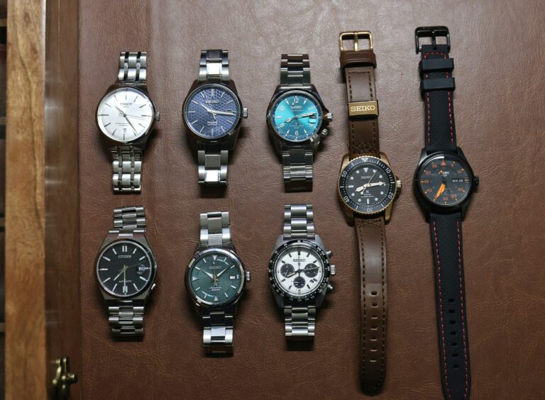 A collection of eight watches on a brown surface, arranged neatly in two rows. Styles vary with metal bracelets and leather straps; tones are diverse.
