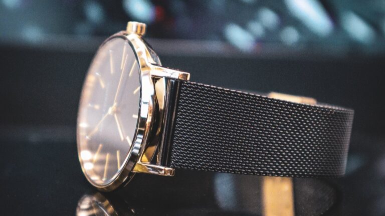 A sleek wristwatch with a golden case, lugs, and a black mesh band rests on a reflective surface.