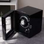 A sleek black watch winder with "FRACO" branding sits open on a gray textured surface, displaying a luxury watch inside.