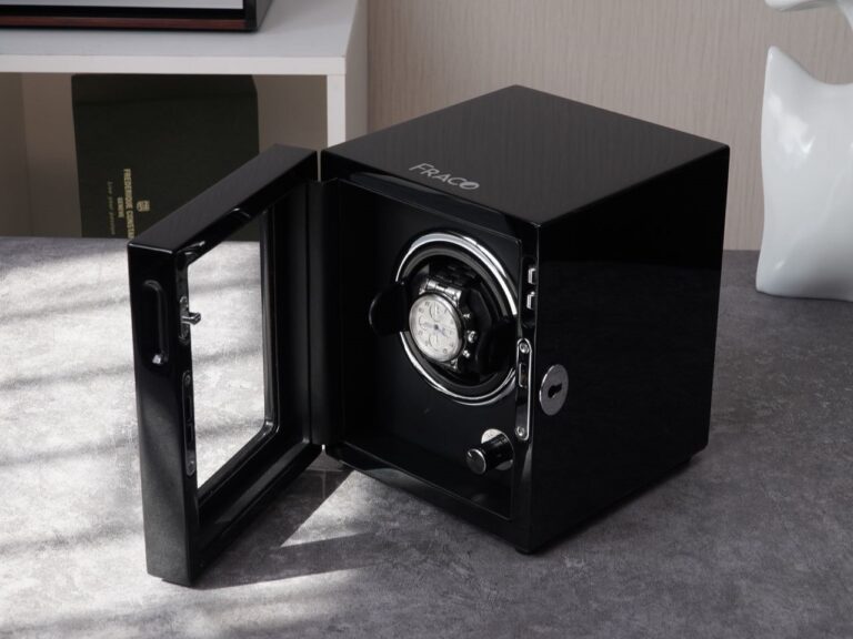A sleek black watch winder with "FRACO" branding sits open on a gray textured surface, displaying a luxury watch inside.